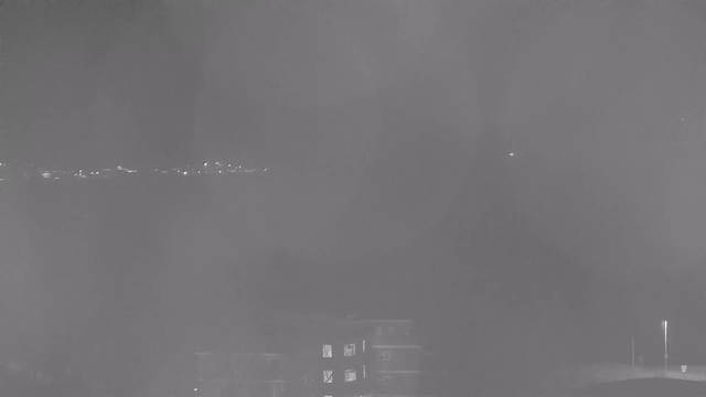 A dimly lit scene taken from a webcam shows a blurred view of distant lights along a shoreline. In the foreground, there is a partially visible building with some illuminated windows. The overall image appears hazy, with a grayish tint, suggesting possible fog or low visibility.