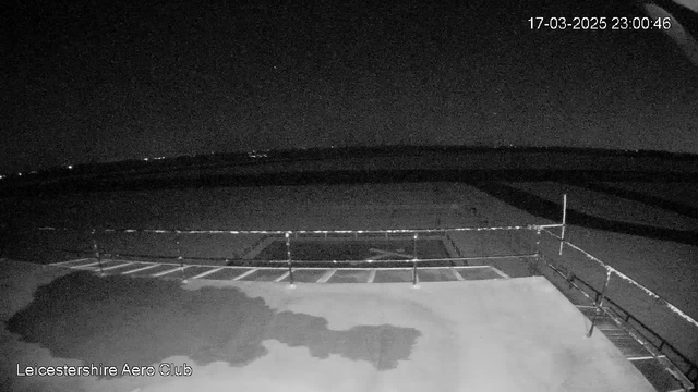 A dark scene captured from a webcam showing a flat surface in the foreground, likely an empty runway or tarmac area at a small airport. Faint lights are visible in the distance, indicating the presence of buildings or vehicles. The sky is mostly dark with a few stars visible, suggesting nighttime. There is a railing along the edge of the tarmac, and the timestamp in the corner indicates the image was taken on March 17, 2025, at 11:00 PM.