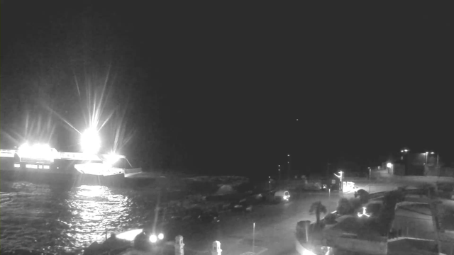 A nighttime scene of a waterfront area. A brightly lit boat is illuminated at the left side of the image, casting reflections on the water. The shoreline is visible with various structures and parked vehicles along the edges. Dim lights from nearby buildings and streetlights can be seen, adding to the ambiance of the area. The overall scene is dark, with the illuminated boat being the most prominent feature.