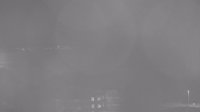 A dimly lit view from a webcam showing a dark, foggy scene. The foreground includes a silhouette of a building with faint lights in the windows. In the background, there is a blurred shoreline with small lights scattered along it, suggesting distant structures or vehicles. The overall image lacks clarity due to the fog and low lighting.