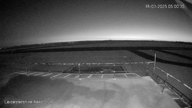 Image of a dark landscape captured at dawn. The foreground shows a flat surface with a rail fence along the edge. There are markings on the ground, indicating possible parking or runway areas. The horizon in the background is gradually lightening, suggesting the sun is about to rise. The sky appears mostly clear with faint outlines of distant landforms. The timestamp indicates the date and time as March 18, 2025, at 5:00 AM.
