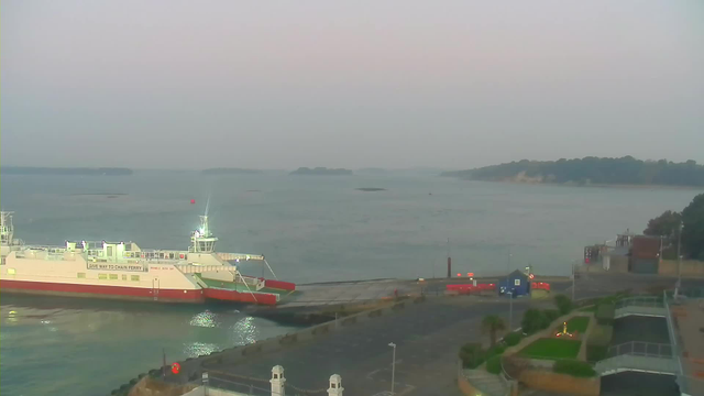 A calm waterfront scene features a large ferry docked at a wharf. The ferry, primarily white with red accents, is illuminated by bright lights. Beyond the ferry, the water is reflective, with several small islands visible in the distance. The sky is soft and pastel-colored, transitioning from light blue to pink hues as the day begins to dawn. In the foreground, there is a neatly arranged area with greenery and pathways, while various structures and vehicles can be seen along the wharf.