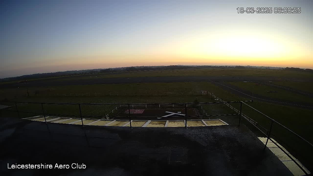 A view from a high vantage point overlooking a long, flat airfield at dawn. The sky is mostly clear, transitioning from deep blue to soft orange near the horizon. A low fence surrounds the airfield, and a small section of grass with a red and white target mark is visible in the foreground. Various asphalt paths and a few markers can be seen on the ground, hinting at the layout of the airstrip. The image is timestamped, indicating it was captured at 6:00 AM on March 18, 2025.
