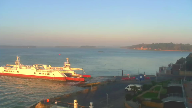 A ferry with a white and red color scheme is docked at a port, with its ramp extended to the shore. The water is calm and reflects the early morning light, while a few small buoys can be seen in the distance. In the background, there are green hills and trees lining the coastline, with a hint of sandy beach visible. Various structures can be seen nearby, including a small blue building and a white van parked on the road. The sky is clear, indicating a bright morning.