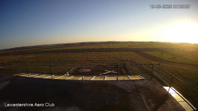 A view from a high vantage point overlooking an open landscape at sunrise. In the foreground, there is a wooden railing enclosing a terrace, with a large white 'X' marking on the ground, possibly indicating a helipad. The ground is mostly grassy with some markings, and a road runs through the scene, leading to the horizon where the sky changes from blue to yellow near the sun. Soft, distant hills can be seen in the background under a clear sky. The time and date are displayed in the upper right corner.