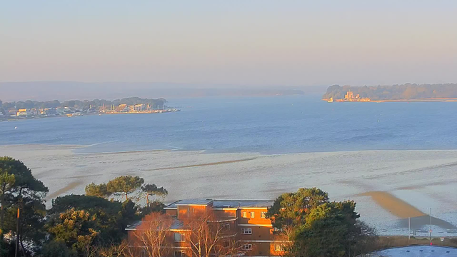 A serene view of a body of water at low tide, with sandy areas visible along the shore. In the background, a town or harbor is seen, featuring various small boats and buildings lining the water's edge. To the right, an island with a structure resembling a castle is visible, surrounded by trees. The sky is clear and slightly hazy, creating a soft glow over the scene. In the foreground, there are patches of greenery, including trees and shrubs, along with the tops of a few buildings.
