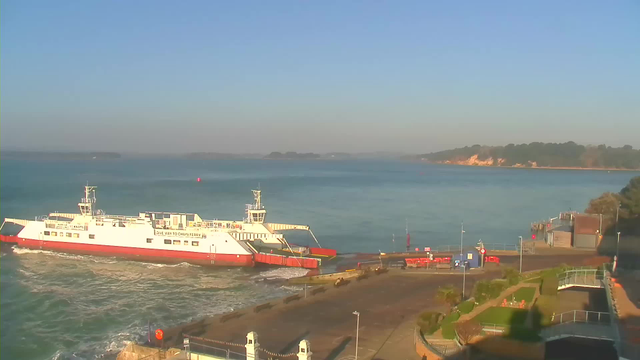 A ferry with a white and red hull is docked at a harbor. The water around the ferry is calm, with gentle waves. The background features a coastline with trees and a sandy shore. In the foreground, there is a paved area with some shrubs and a small garden. Several posts and signs are visible, and there are a few vehicles parked nearby. The sky is clear and sunny.
