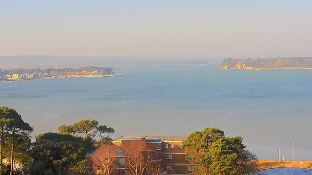 A serene view of a calm body of water, with gentle waves reflecting the soft blue sky. In the background, a coastal area features several boats moored at a marina, alongside residential buildings. Dense trees frame the lower part of the image, and a distant shore with land formations is visible on the right side. The overall atmosphere is tranquil and picturesque, suggesting a peaceful seaside location.