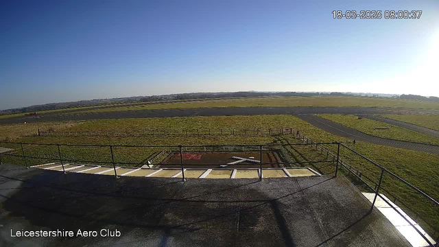 A clear blue sky stretches above a wide, open grassy field connected to a runway. In the foreground, a terrace with a railing is visible, leading to a helipad marked with a large white "X". The surrounding area features markings on the runway and a perimeter fence, with distant hills visible beyond the field. The scene is bright and sunny, indicating a clear day.