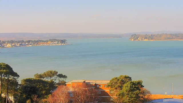 A view of a calm blue water body that stretches into the distance, bordered by trees and land on either side. In the foreground, there is a multi-story brick building among vegetation. On the left, a small town or marina with boats can be seen along the shore, while on the right, an island with structures is partially visible. The sky above is bright and clear, suggesting a sunny day.