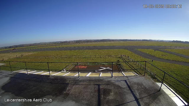 A view from a webcam showing a clear blue sky above an expansive green field with a runway. In the foreground, there is a ground-level structure with a metal railing. A white cross is visible on the ground, indicating a helicopter landing area. Several wooden fences line the edges of the grassy area.