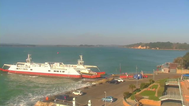 A ferry docked at a harbor on the coast, surrounded by calm blue water. The ferry is white with red accents, and there are several vehicles parked nearby on the shore. In the background, a coastal landscape features green hills and a sandy cliff. The sky is clear and sunny, creating a bright atmosphere.