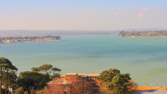 A scenic view of a tranquil body of water, showcasing shades of blue and green. In the foreground, there's a reddish-brown building surrounded by trees. To the left, a marina filled with boats is visible, leading to a distant shoreline with more trees and buildings. The sky is mostly clear with a few clouds, and the landscape fades into gentle hills in the background.