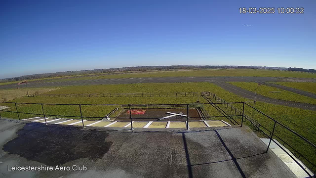 A clear blue sky fills the upper portion of the image. Below, a large, open grassy area stretches out, intersected by two runways made of dark asphalt. In the foreground, there is a railing overlooking a helipad with white markings forming a cross and another red area. The scene suggests a wide, unobstructed aviation field with a distant horizon, indicating a bright, sunny day.