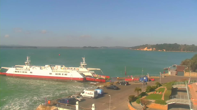 A bright day at a harbor with a turquoise sea. In the foreground, a white and red ferry is docked, with its loading ramp lowered. Nearby, several vehicles are parked, including cars and a van. The area features green grass, landscaping, and pathways. In the background, a sandy shoreline with trees and cliffs stretches along the coast. The sky is mostly clear with a few clouds.
