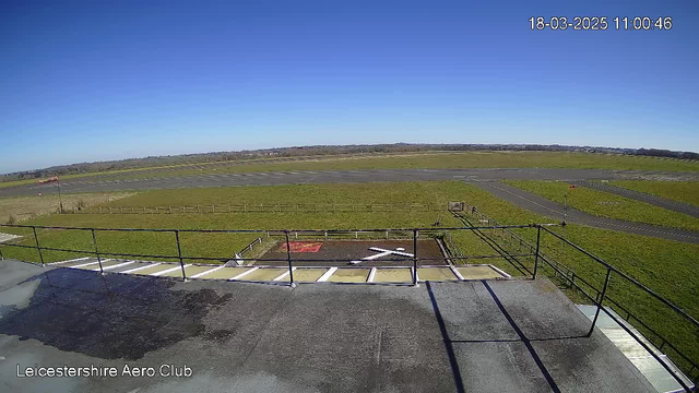 A clear blue sky stretches overhead, with no clouds visible. The foreground shows a flat rooftop with black and white stripes, and a railing along its edge. Below, an open field is viewed, with a well-maintained grassy area, a runway paved in gray, and a visible taxiway. In the distance, there are distant hills and a few scattered trees. A windsock is seen at the edge, indicating wind direction. The scene is tranquil, typical for an aerodrome setting.
