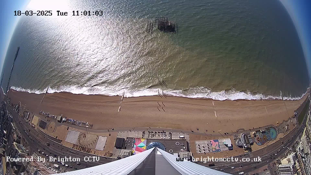 A bird's-eye view of a sandy beach with gentle waves lapping at the shore. In the distance, an old pier extends into the water. The beach is lined with colorful beach huts and a carnival area featuring rides and games. People are walking along the beach and the promenade, and the sky is clear with sunlight reflecting off the ocean's surface. The image contains time and source details in the corners.