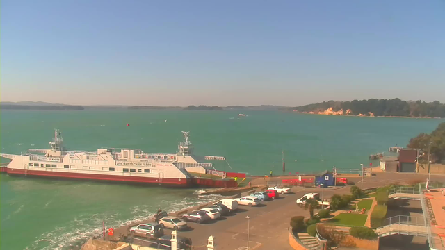 A sunny waterfront scene featuring a large ferry docked at a pier. The ferry is white with red accents and has signage indicating it is a chain ferry. The water is a bright turquoise color, with gentle waves visible. In the foreground, there is a parking area with several parked cars, and a pathway lined with greenery and trees. On the right side, steps lead down from a raised area towards the water. In the background, a hilly shoreline is visible with trees and rocky cliffs. The sky is clear with a few distant hills on the horizon.