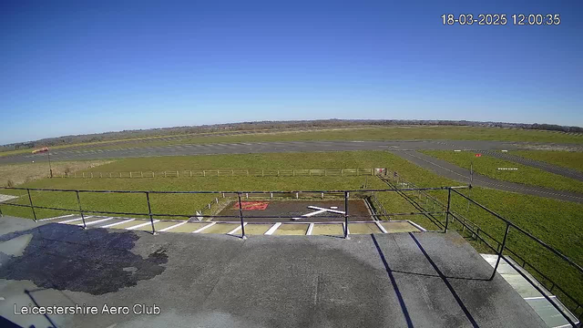 A clear blue sky stretches above a grassy airfield, with a runway extending into the distance. In the foreground, there is a flat rooftop with a railing, and a marked helipad with an "X" shape. The airfield features patches of green grass and a few dirt sections, and there are faint lines marking the edges of the runway. The overall scene is bright and open.