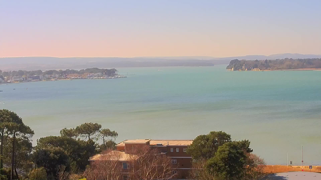 A serene waterfront scene featuring a calm body of water in shades of blue and green. In the background, trees line the shore, and several buildings are visible along the waterfront. Some boats are scattered on the water, and distant hills create a soft horizon. A foreground of greenery includes trees and shrubs, while a brick building is partially visible at the bottom of the image. The sky transitions from light blue to a soft pink near the horizon, suggesting either early morning or late afternoon light.