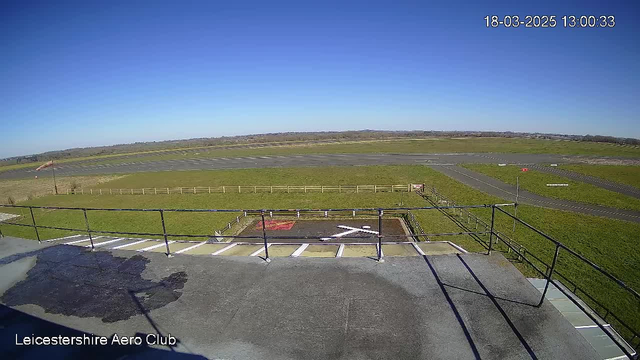 A clear blue sky is visible above a large open area of grass and paved runway at the Leicestershire Aero Club. In the foreground, there is a railing and a flat rooftop surface with an "X" marking on it, indicating a landing zone. Several grassy patches are visible on the ground, and a fence outlines the aerodrome's perimeter. The scene is bright and sunny, indicating a clear day. A small windsock is present, showing the wind direction.