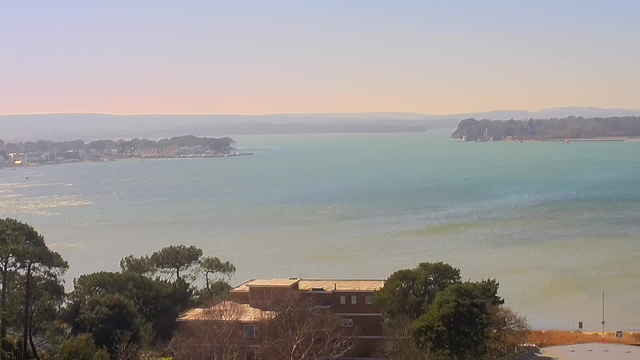 A scenic view of a calm body of water, with soft blue and green hues reflecting the sunlight. In the foreground, there are trees and a brown building. The background shows a marina with several boats, gently bobbing on the water. Hills and trees are visible in the distance, creating a serene landscape under a clear sky.