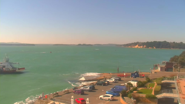 A view of a turquoise sea under a clear sky. In the foreground, there is a parking area with various vehicles, including a red car. On the right, a path leads to a waterfront area with greenery and structures. In the distance, there are small islands and a coastline with sandy cliffs. Waves gently lap at the shore, indicating calm weather.