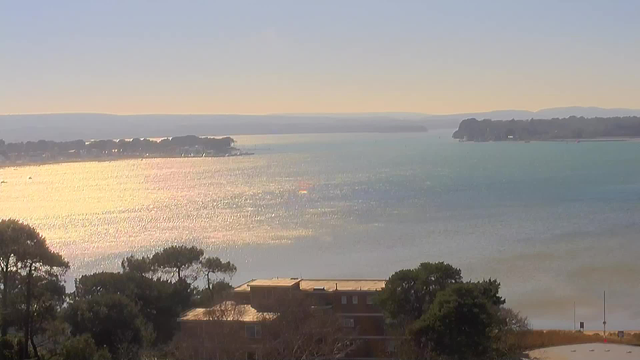 A scenic view of a body of water reflecting sunlight, with light blue and turquoise hues. In the foreground, there are trees and some buildings partially visible. The background features a distant shoreline with more trees and hills under a clear sky. The overall atmosphere is calm and serene, capturing the beauty of nature.