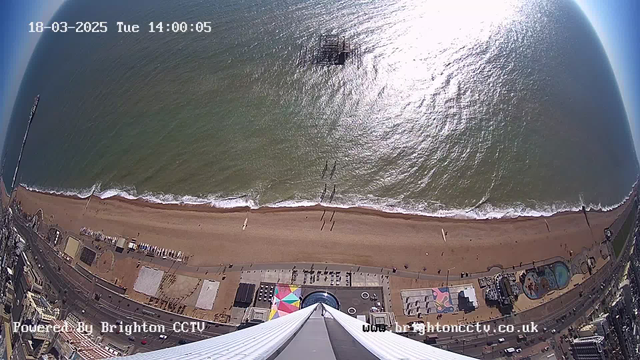 A view from above a beach, showing golden sands and gentle waves lapping at the shore. People can be seen walking on the beach, and a pier extends into the water. There are various colorful structures and attractions along the beachfront, including amusement rides and a pool area. The sky is clear, and the sunlight creates a shimmering effect on the water's surface. The scene is serene, capturing the essence of a sunny day at the seaside.