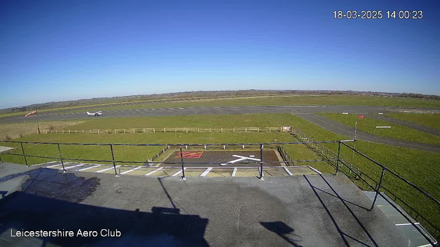 A clear blue sky occupies the background of the image, with a small airplane taxiing on a runway that stretches horizontally across the center. In the foreground, there is a railing and a flat surface, suggesting a viewing platform. To the left, a windsock sways gently, indicating wind direction. The runway is bordered by green grass on both sides, with a fence visible in the distance. Overall, the scene depicts a sunny day at an airfield, with the airplane in motion.