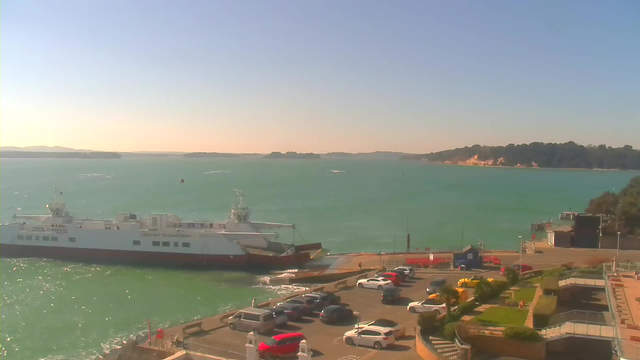 The image shows a tranquil waterfront scene with a large ferry docked at a pier. The ferry has a white and red color scheme and appears to be waiting to board passengers. In the background, the water is a bright turquoise, with small waves gently rippling. Beyond the water, there are distant hills and a sandy shoreline with trees. In the foreground, there’s a parking lot filled with vehicles of various colors, including white, red, and dark blue, along with some greenery and a pathway leading to the pier. The sky is clear and bright, indicating a sunny day.