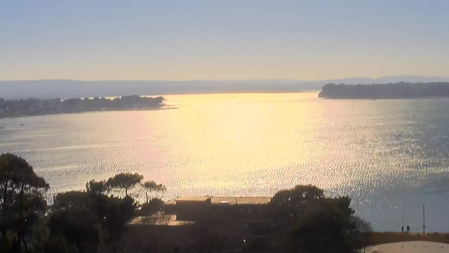 A serene view of a sparkling body of water reflecting sunlight, creating shimmering golden and silvery patterns on the surface. In the background, there are distant hills and trees along the shoreline. The sky above is clear with hints of soft colors, indicating early morning or late afternoon light.