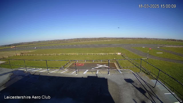 A clear blue sky is visible above a grassy airfield. In the foreground, there is a railing along the edge of a building. Below the railing, a helipad with a red 'X' marking is present. A small white aircraft is positioned on the runway to the right, while a few parked vehicles can be seen in the distance. The airfield is surrounded by a wooden fence, and the landscape extends towards the horizon, showing fields and distant trees.