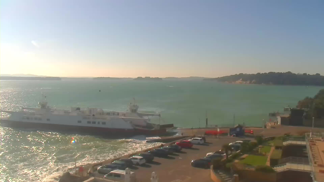 A coastal view featuring a large ferry docked at a pier. The water is turquoise and has gentle waves, reflecting sunlight. In the foreground, there is a parking area with several cars, and to the right, some greenery and modern buildings can be seen. The sky is clear with a few wispy clouds, and distant hills are visible on the horizon.