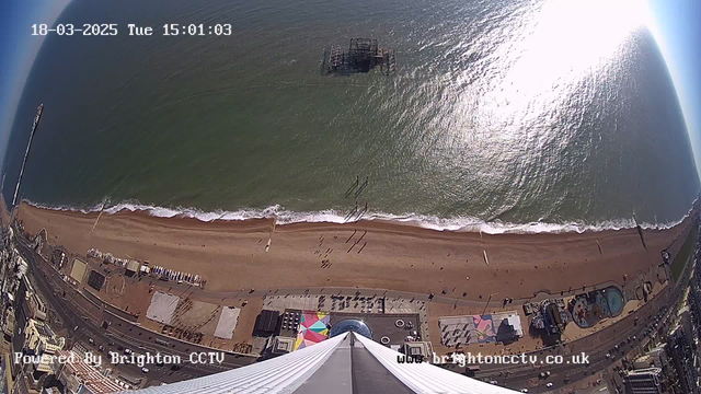 A high-angle view of a beach scene taken from a webcam, showing a sandy beach with people walking along the shore. The ocean waves gently roll in, reflecting sunlight. In the background, a pier with a structure can be seen, while colorful beach huts and attractions are visible along the beachfront. The image is timestamped at 15:01 on March 18, 2025.