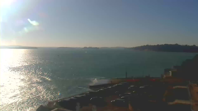 A bright, sunny day with a clear blue sky and a shimmering ocean. The water reflects sunlight, creating a sparkling effect on the surface. In the foreground, there is a parking area with several cars, and to the right, a pier or walkway leading toward the water. Distant landforms can be seen on the horizon, suggesting islands or the coastline.