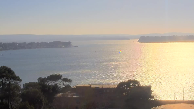 A wide view of a calm body of water reflecting golden light from the sun. In the distance, there are hills and trees lining the shore. Several small boats can be seen on the water. The sky above is clear with a soft gradient from light blue at the horizon to a lighter color near the sun. The shoreline features partial buildings and vegetation.