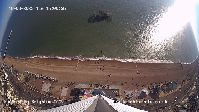 A high vantage point view of a beach with a sandy shoreline and gentle waves lapping at the edge. In the background, the water is shimmering under sunlight, reflecting light. A pier extends into the water, and a large structure, possibly a pier or amusement park, is visible off the shore. People can be seen walking along the beach, and various buildings and colorful attractions are located near the beach area. The image also includes a timestamp indicating the date and time.