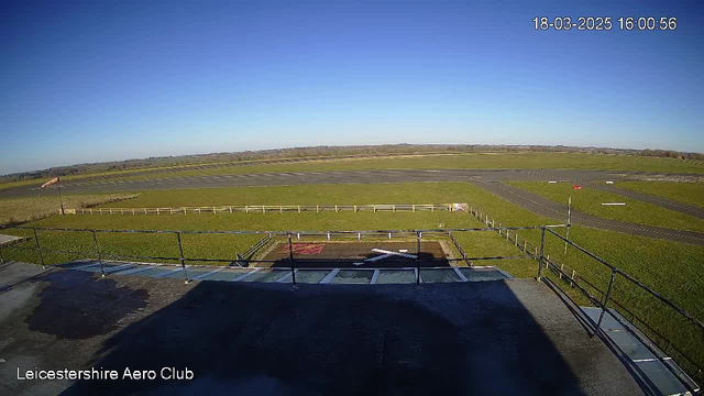 A clear blue sky is visible, with vast green fields stretching across the scene. In the foreground, there is a flat rooftop with a railing, partially shaded by its own structure. A grassy area with a white fence can be seen beyond the rooftop, leading to an asphalt runway. Along the runway, there are several markings and a small red flag indicating wind direction. The overall atmosphere is bright and open, typical of an airfield setting.
