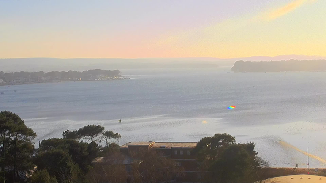 A scenic view of a calm body of water reflecting soft pastel colors from the sky, where the sun is low on the horizon. In the distance, there are trees lining the shore, and a few small boats are visible on the water. In the foreground, there are some trees and buildings, adding depth to the landscape. The image conveys a serene atmosphere with gentle ripples on the water's surface.