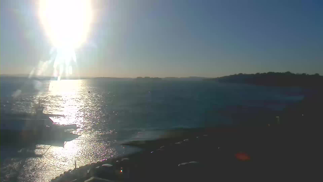 A bright sun shines in the sky, creating a glare on the water below. The surface of the water reflects the sunlight, appearing sparkling and shimmering. In the foreground, a silhouette of a boat is visible, while the background features a distant shoreline with hills and trees. The sky is clear with no visible clouds, transitioning from blue near the horizon to a lighter shade above the sun.