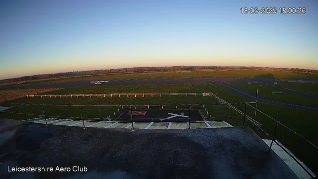 A clear blue sky stretches overhead, with the sun low on the horizon. Below, an expansive green field meets a tarmacked runway at an airport. In the foreground, there is a low railing and a flat rooftop area. A white airplane is parked on the runway, while a few light-colored markers line the field and runway. The scene is serene, indicating a late afternoon at an airport, with no visible activity.