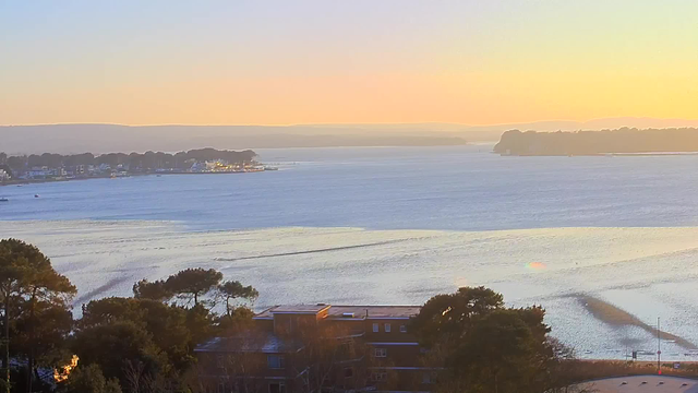 A tranquil view of a body of water during sunset, with soft pastel colors in the sky. In the foreground, there are trees and the top of a building with a flat roof. The water features gentle ripples, and in the distance, there are small boats and buildings along a shoreline that is partially obscured by trees. A line of hills is visible beyond the water, creating a serene backdrop.