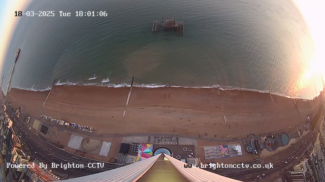 A bird's-eye view of a sandy beach with people walking along the shore. The water is calm with small waves lapping at the beach. In the background, a pier extends into the water, featuring a structure that appears to be partially submerged. The coastline is lined with buildings and attractions, including a fairground, visible near the lower part of the image. The scene is bathed in the warm glow of the setting sun. The date and time are displayed at the top, indicating March 18, 2025, at 6:01 PM.