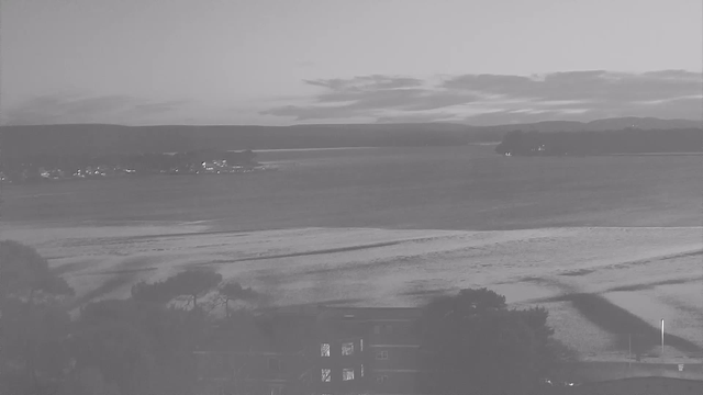A monochrome image of a tranquil water scene, featuring a wide body of water reflecting soft light. In the background, there are distant hills under a cloudy sky. Near the water's edge, sandy patches can be seen, with some trees framing the lower part of the image. A building is partially visible in the foreground, hinting at its presence near the shore. The overall atmosphere conveys serenity and calmness.