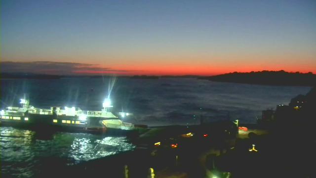 A twilight scene featuring a large ferry docked at a terminal surrounded by water. The sky is transitioning from blue to deep orange and red near the horizon. The water reflects the fading light, creating a shimmering effect. There are faint outlines of distant hills and a few lights from vehicles and buildings along the shore.