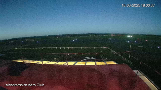 A view from a webcam shows a darkening sky at dusk over an open field, with faint lights scattered throughout. In the foreground, there is a flat red-roofed structure, with a ledge or railing. The field beyond is mostly empty, but features a white fence outlining an area, possibly for aviation activities. The overall mood is tranquil, with a hint of evening activity indicated by the lights in the distance.
