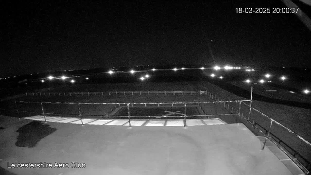 A dimly lit scene from a webcam, showing a large, flat rooftop in the foreground with a horizontal railing. In the background, there are rows of lights in the distance, suggesting an airfield or similar open area, under a dark night sky. A few scattered clouds may be visible, but overall the image is predominantly dark. The time displayed in the top right corner reads 20:00:37 on March 18, 2025.