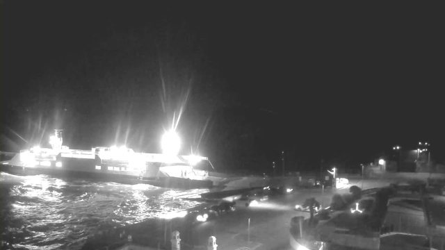 A brightly lit ferry is docked at a port at night, with strong lights illuminating the boat. The water is choppy and reflects the lights. Several vehicles are positioned near the ferry, and there are faint outlines of structures and lights in the background, indicating a busy harbor setting.