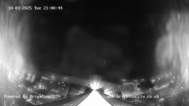 A black and white image from a webcam showing a dark urban scene at night. The center features a bright light source, possibly a streetlight or building interior, creating a glimmering effect. The edges are more shadowy, with faint outlines of structures and lights. There is text in the lower left corner that reads "Powered By Brighton CCTV" and a timestamp at the top indicating the date and time.